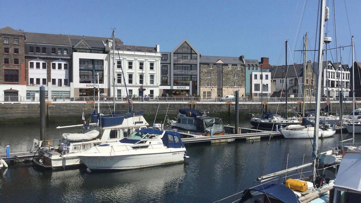 A view from the other side of the Quay, which shows a set of modern and old buildings next to each other, some with large windows, others with a stone exterior, and another with an old white exterior. There are boats in the harbour in the foreground.