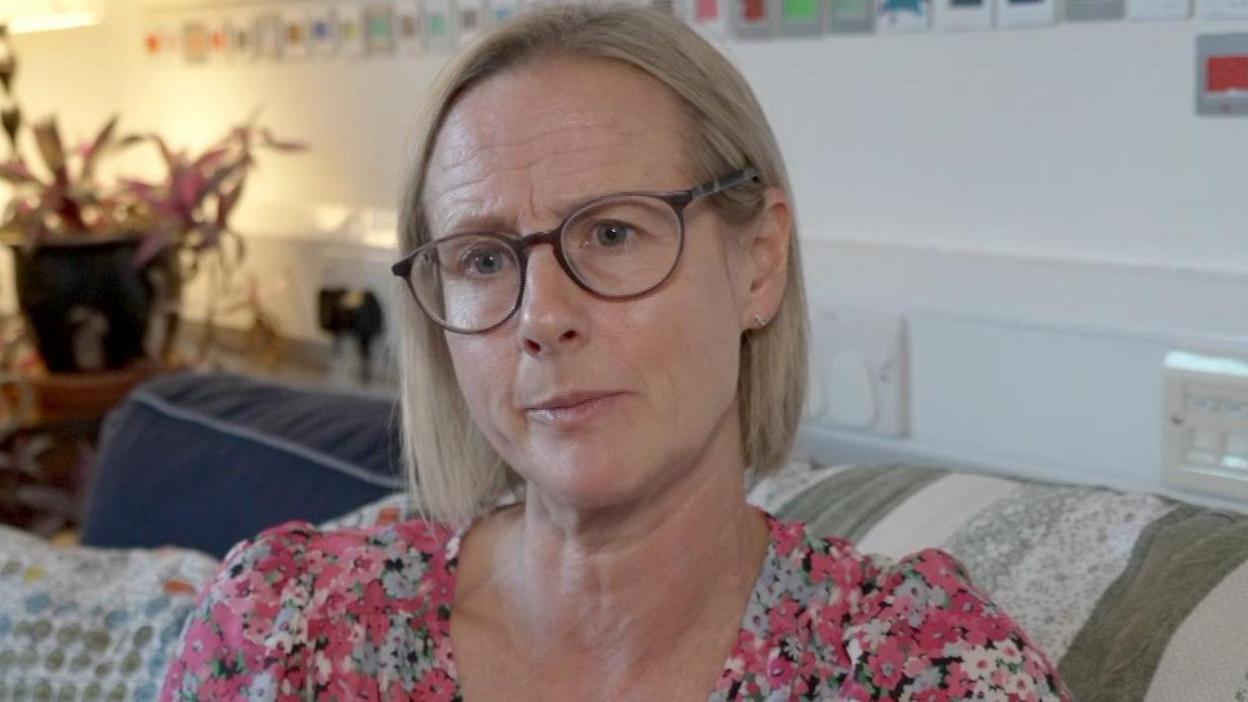 Heather Richards has short blonde hair and round glasses. She is wearing a pink floral top.