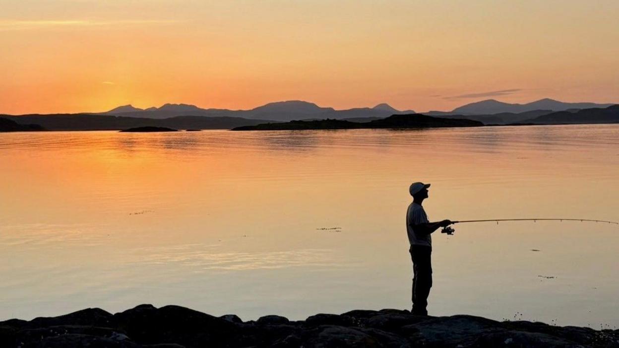 Silhouette of a person wearing a baseball cap and holding a fishing rod standing on the beach as the sun sets over a body of water behind them and hills in the distance