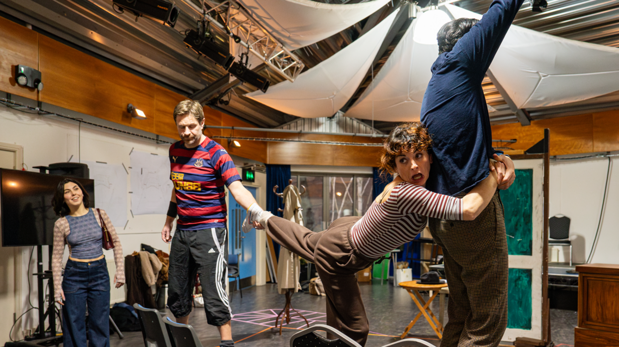 Four actors in a large rehearsal room with a rough scene set up including a door, a coat stand and four dining room chairs. Three of them are standing on the chairs with one reaching upwards and another clinging on to his waist with her leg being pulled by another man