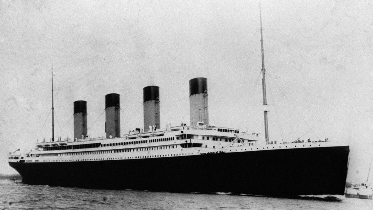 A black and white image of the Titanic, a large ship with a black hull and white cabins with four large funnels