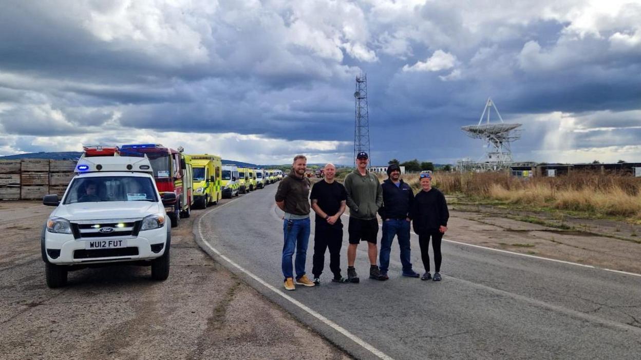 Five people stood on a road beside a row of parked emergency vehicles, with a large satellite dish on the horizon