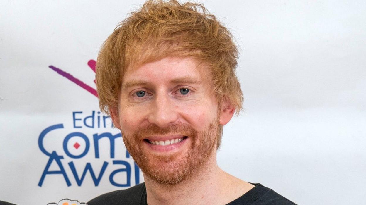 Mark Simmons on close up, smiling to camera. An Edinburgh Comedy Award banner can be seen behind him.