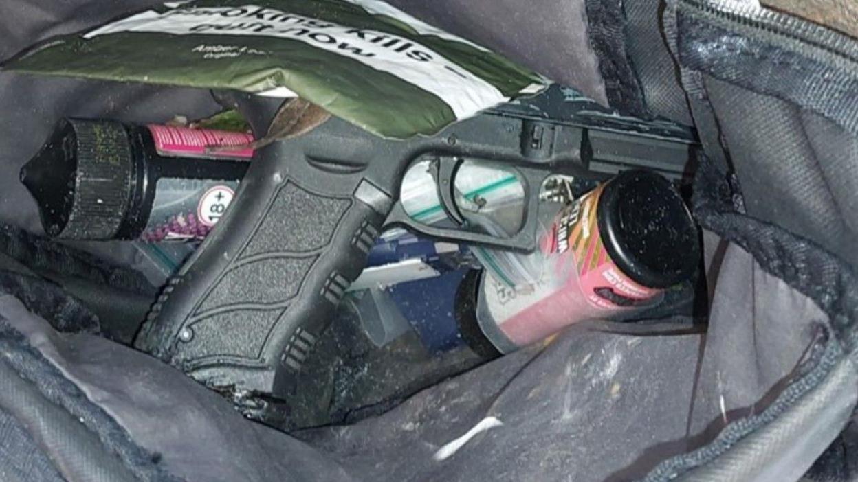 A gun near other items. The gun is black and rests in a bag on top of a can and a small plastic bag with another bag with the words "smoking kills" resting on top of it.