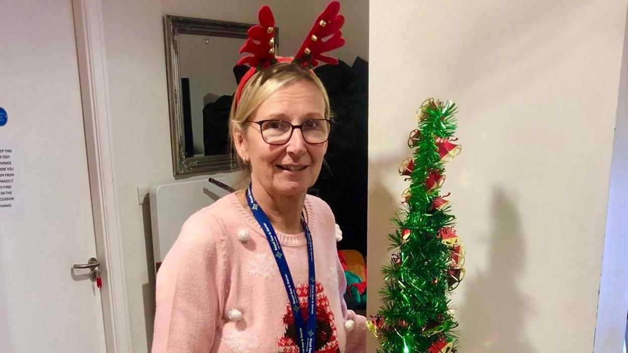 A blonde haired woman wearing a pink jumper, reindeer ears, and glasses has a staff lanyard on, as she looks at the camera with a small Christmas tree next to her 