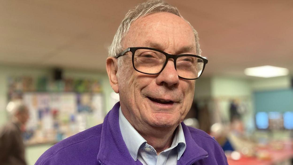 A close-up of Tony smiling at camera, wearing a purple fleece with the BDAA logo on it and with the memory cafe taking place behind in a blurred background