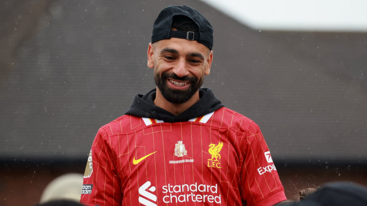 Mohammed Salah is wearing a red Liverpool football shirt. He has a black cap on backwards and is smiling. He has a black beard and moustache.