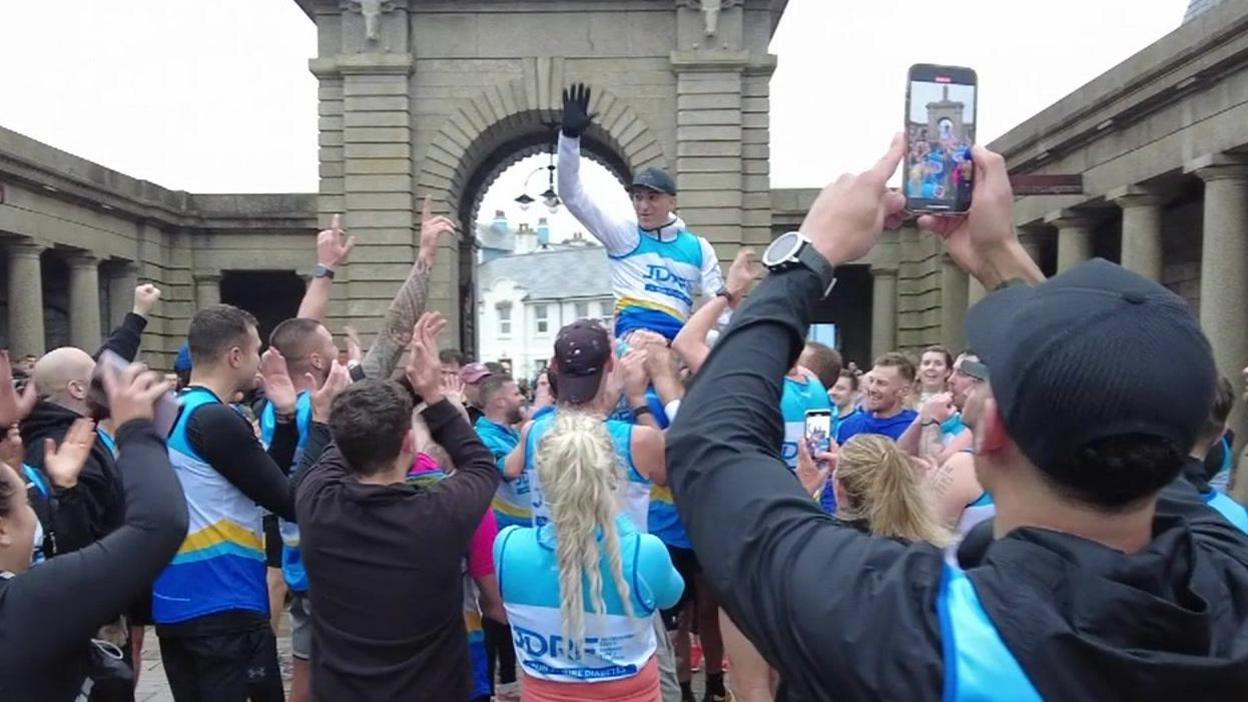 Mehdi Taheri smiles and waves at a crowd of people he has been hoisted up in the middle of. He is wearing a white and blue running kit. People have their hands up in the air and are clapping him outside Royal William Yard.