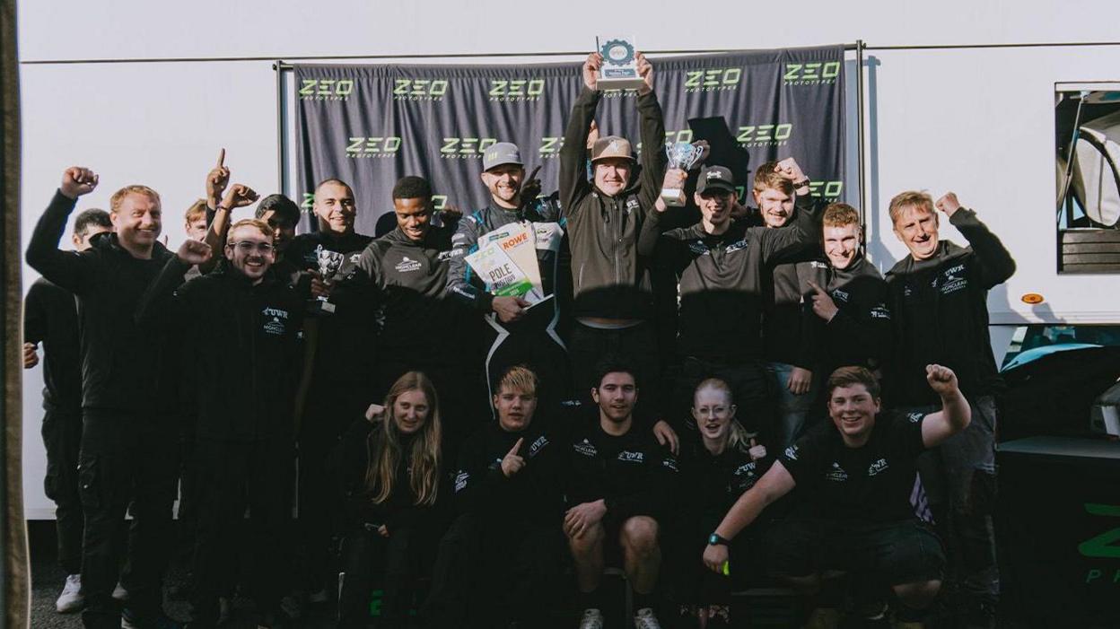 A racing team dressed in black pose for a team picture, with some raising their hands and one holding up a trophy. They are standing in front of a black banner that says "ZEO" on it in yellow/green letters.