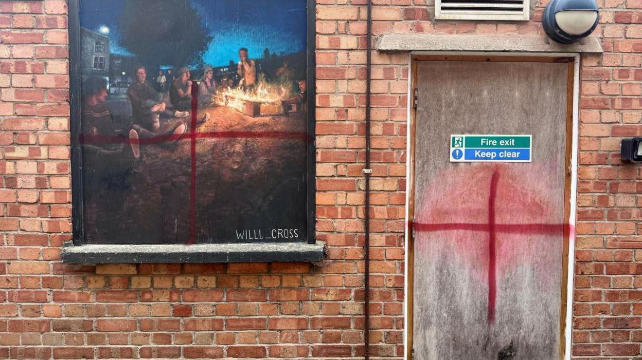 Two painted crosses - one on a grey fire exit door and another over an ornate mural showing people singing around a fire. Both are set into a red brick wall. 