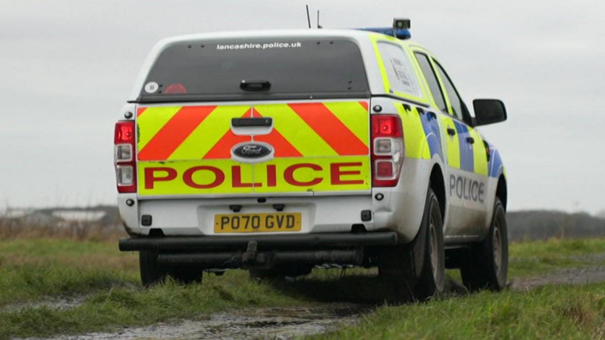 liveried police car driving along a dirt track on a field, lined with grass