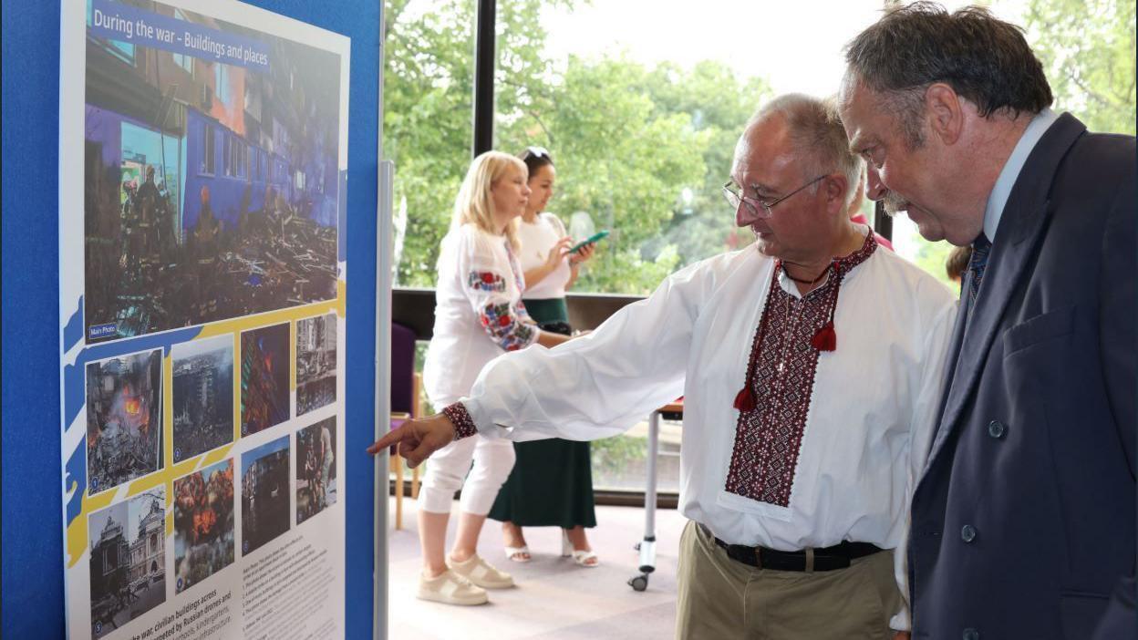 Two men standing in front of a board surrounded by a blue and yellow border covered in photographs of Ukraine and Ukrainian refugees
