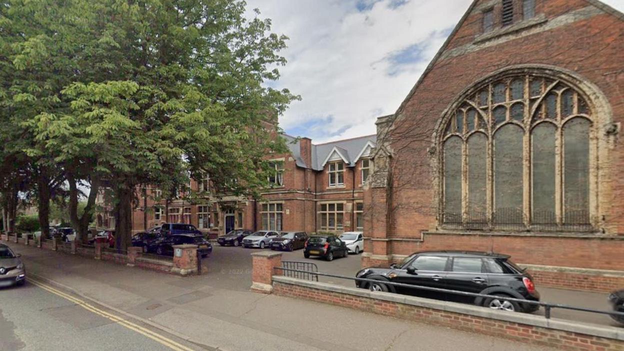 An exterior view of the King's (Cathedral) School in Peterborough. It shows bred brick Victorian or Edwardian buildings with large windows, with a low wall along a wide pavement. There is a gateway for cars in the wall and many cars are parked on the tarmac. There are large trees in leaf shading the tarmac in the top left.