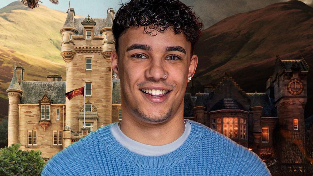 Freddie, a 20-year-old man, who has short curly hair and stubble and is smiling broadly. He is wearing studs in his ears and a blue crew neck jumper over a paler blue t-shirt. Behind him are hills rising to the sky and a stone Scottish baronial-style castle with creamy stone, a grey slate roof and turrets