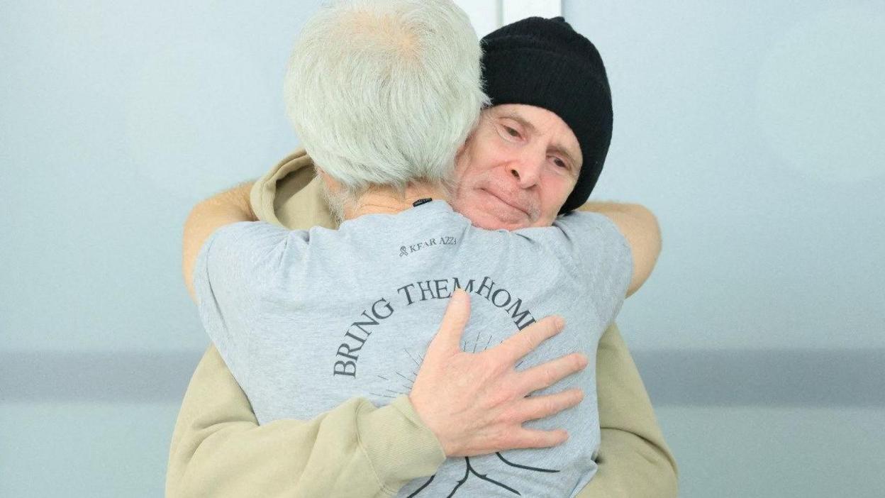 Released American-Israeli hostage, Keith Siegel, wearing a hat, embraces a loved one, the back of whose shirt reads "Bring them home" in an Israeli hospital