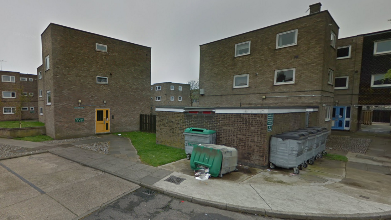 Blocks of pale grey brick three-storey flats, with white window frames. Industrial wheelie bins are in the foreground, with one knocked over 