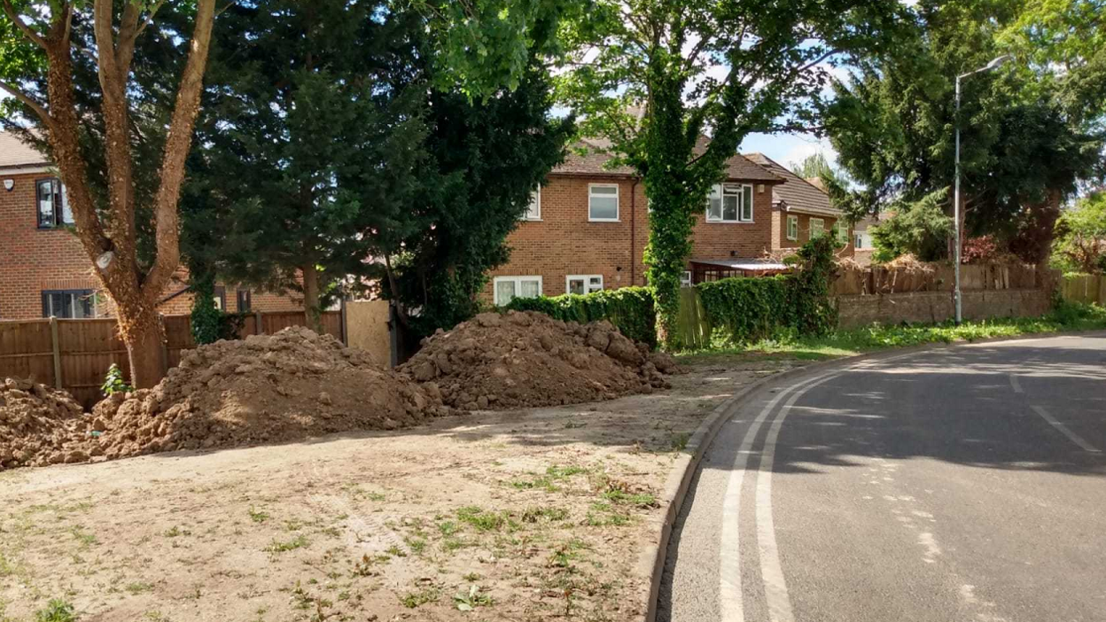 A picture taken aside a road, on the right, and very dry land, with three large piles of dug-up soil, on the left. There are sprigs of grass sprouting from the land. There are homes, which are fenced off, behind the land.