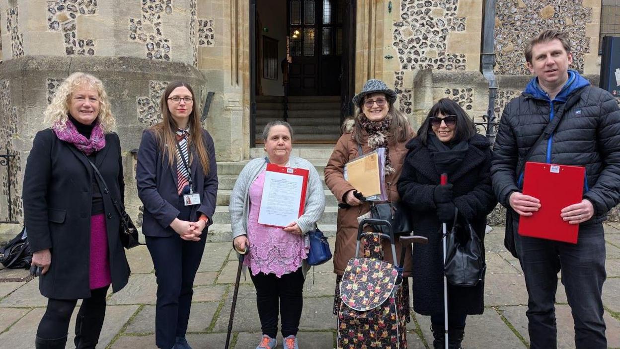 A group of six people stand in front of a stone-walled building holding clipboards containing signatures on a petition. The group is made up of 5 women and one man. The two people on the left of the image are Councillor Lulu Bowerman and Councillor Kirsty North.