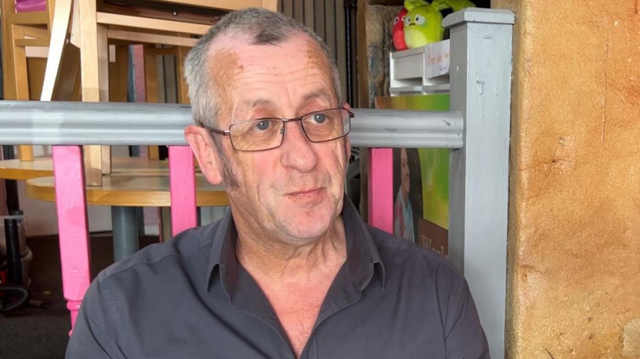 Tony MacDonald has grey hair and sideburns and is wearing thin-rimmed glasses. He is pictured seated in the cafe where he has delivered the training courses and is wearing a dark grey shirt.