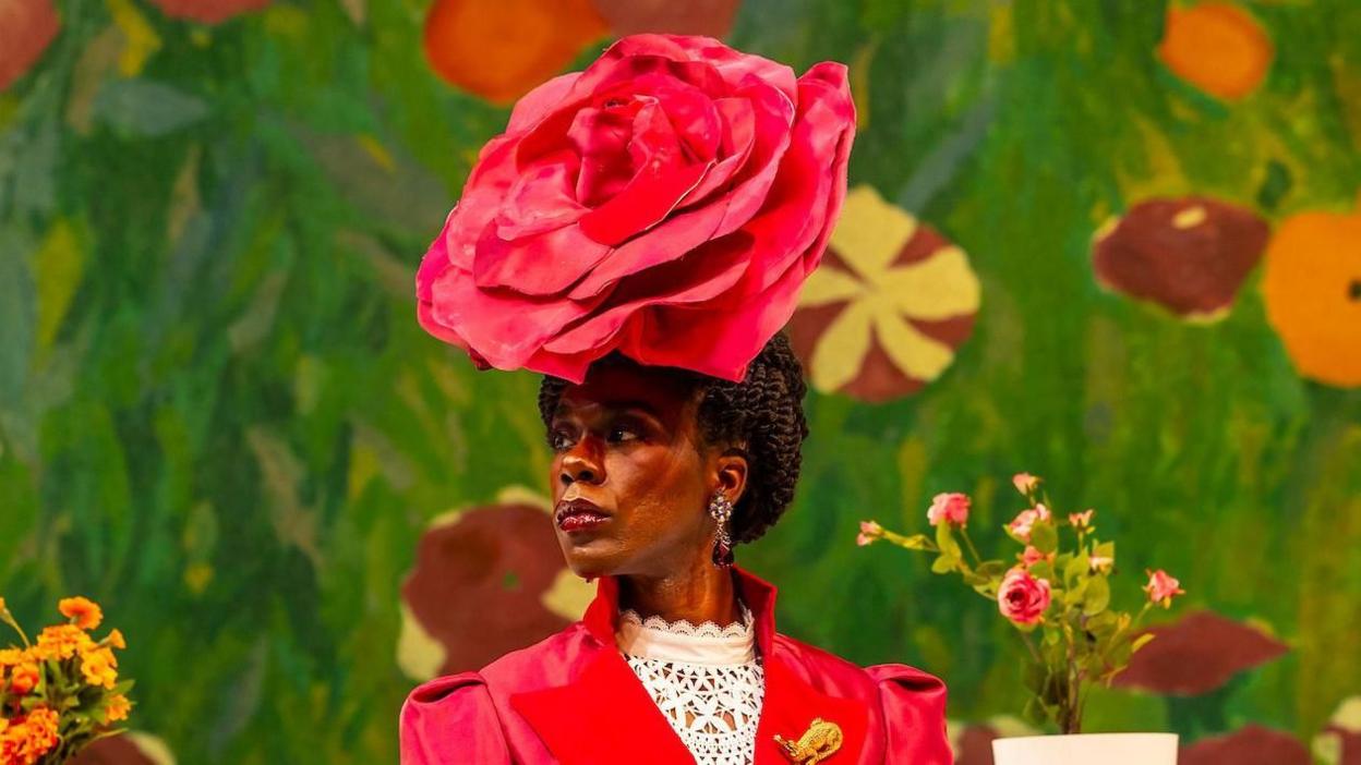 Kajoba dressed as Lady Augusta Bracknell on stage. He looks away from the camera. He wears a large pink headdress in the shape of a rose. He wears a pink dress with a white blouse underneath.  