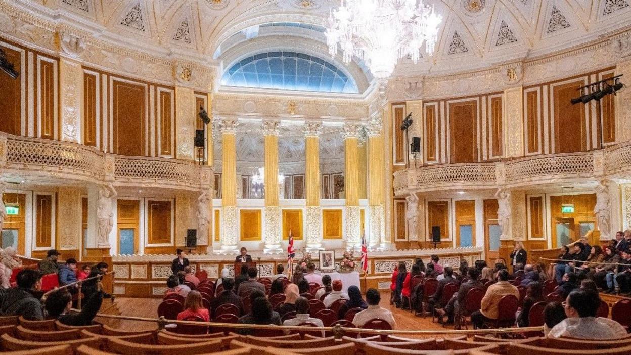 Rows of people of all ages are seated inside St George's Hall, with the historic surroundings including a large chandelier. A number of British flags and a framed photo of King Charles can be seen at the front of the hall.