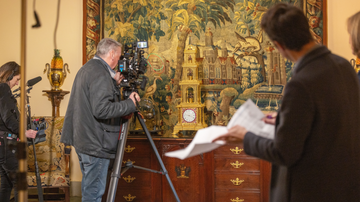 A film crew inside the Abbey looking at a detailed tapestry of birds and trees. A man with a camera on a tripod is stood in front of a wooden set of draws to film the art. A woman with a boom microphone stand behind him.