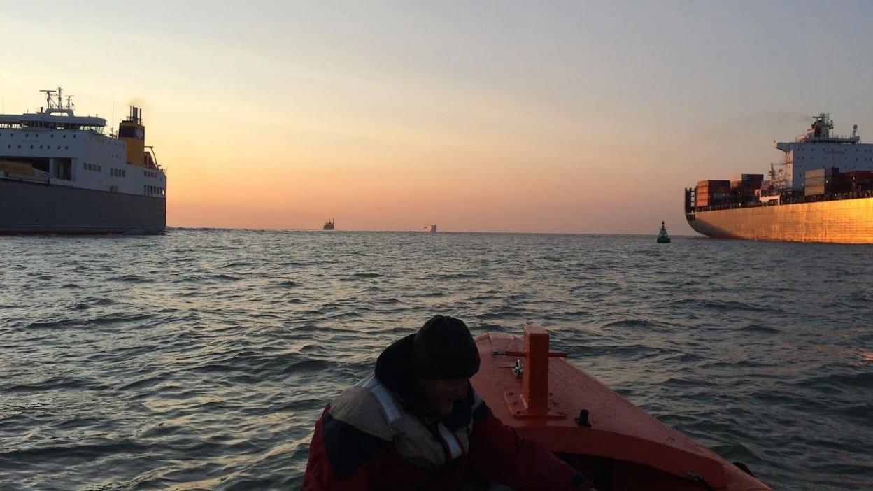 A man on a dinghy in the foreground with two large container ships on either side and two container ships in the distance, the Thames Estuary 