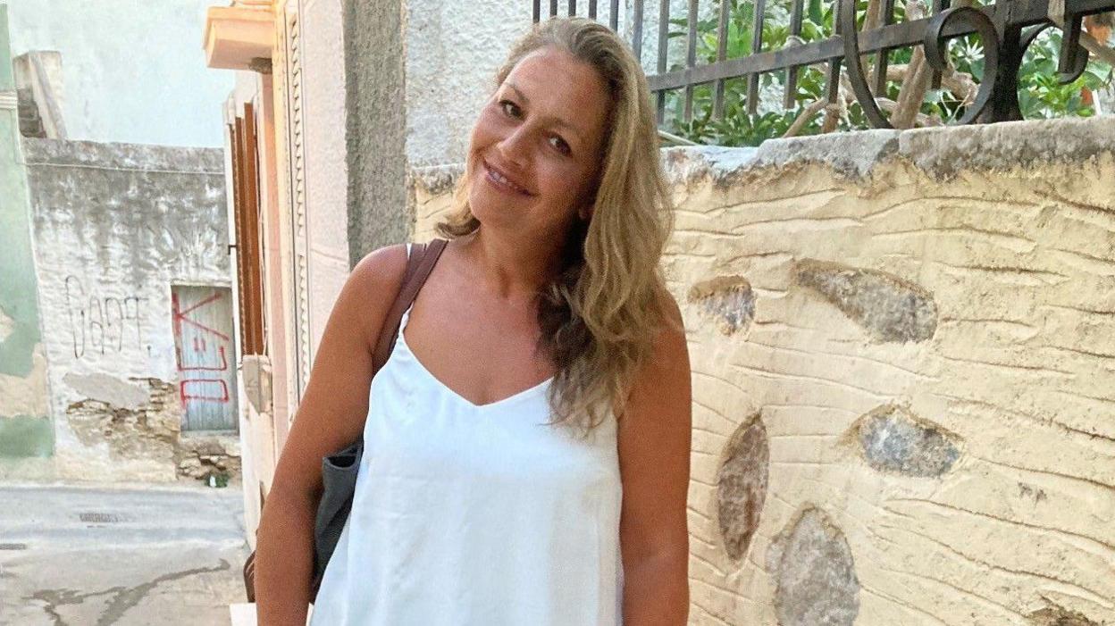 Victoria is standing by a stone wall on a Greek street, with blonde hair and wearing a white vest top.