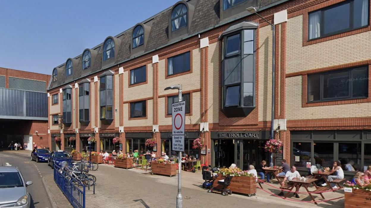 The Troll Cart is a brick building with several windows, outside of which people can be seen sitting in the sun at tables, eating and drinking. A small taxi rank can be seen on the left of the image