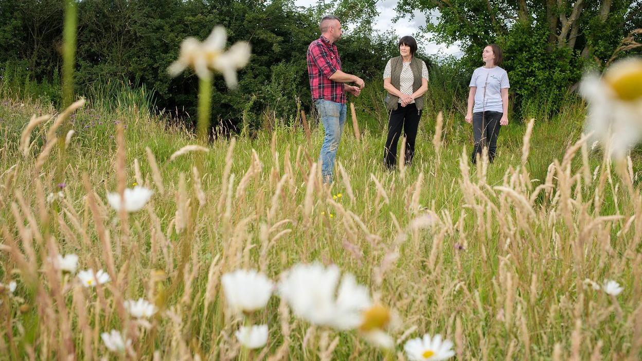 Grass with white flowers in the foreground. Will, is who wearing a red and black shirt with blue jeans, is standing next to Kim - who is wearing a white T-shirt, green gilet and dark trousers - and Hannah, who is in a light-coloured T-shirt and dark trousers, in the distance. There are trees and bushes behind them.