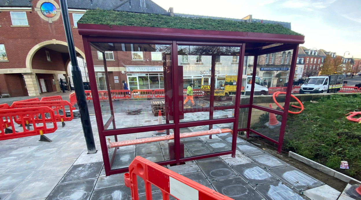 A new bus shelter in the town on freshly-laid paving slabs and surrounded by red barrier gates and a workman in a high vis jacket in the background