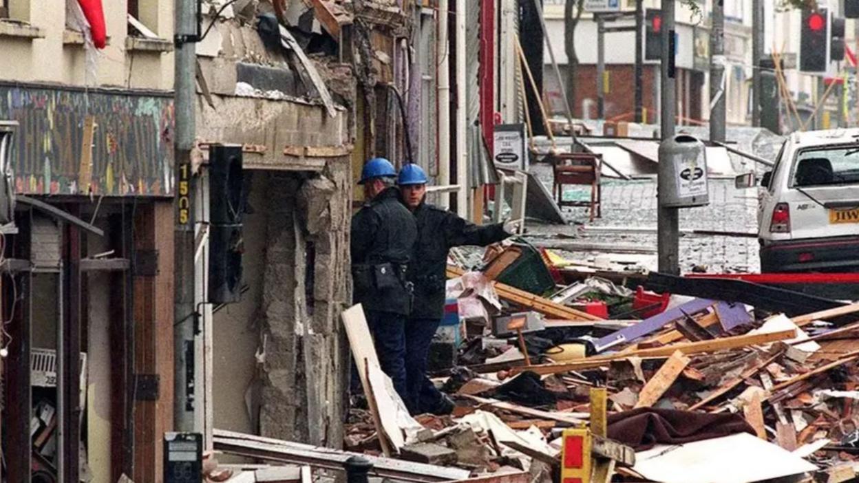 Scene of the Omagh bombing. Two men wearing blue overalls and blue hard hats survey the destruction.