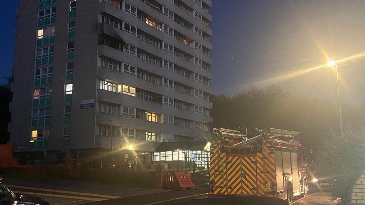 Wider shot of Longfield house, showing a fire engine outside and lights glowing in the dusk