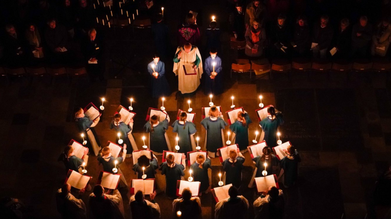 An aerial shot from high in Salisbury Cathedral looking down in darkness at a choir with each singer holding a lighted candle, looking out towards the congregation.