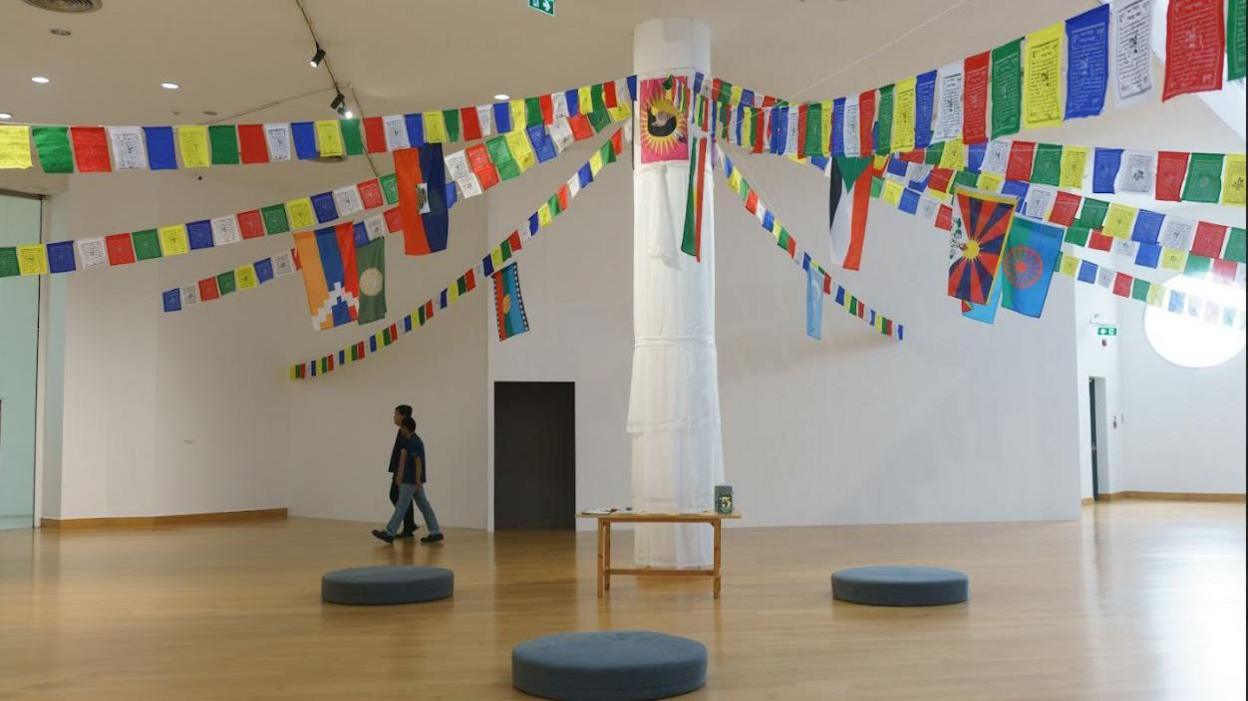 A large pillar stands in the middle of a gallery wrapped in white cloth, with several strings of colourful bunting carrying the flags of various countries and Tibetan art tied to the top of the pillar where a red and yellow flag has been pinned. There is a small wooden table at the base of the pillar as well as black circular cushions placed around it. Two people dressed in jeans and dark T-shirts are walking across the gallery