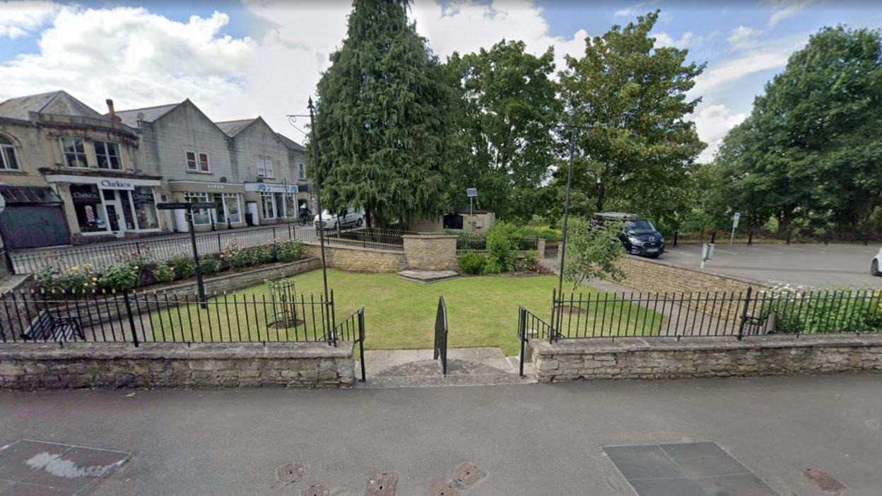 Queen Mary Garden. It is a triangular shaped grassy patch in the middle of a town square. There is a stone wall and iron railings around its circumference and several shops to its left and a carpark to its right. 