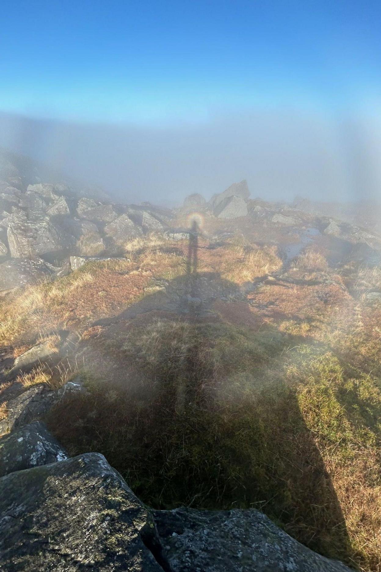 Milo's shadow on low cloud is surrounded by a halo. The shadow has formed in a mountain landscape of large rocks.