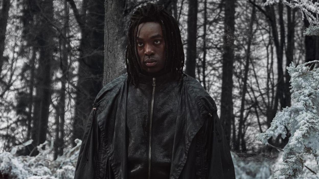 Paapa Essiedu in a black zip-up jacket and cape looking moody in a snowy forest