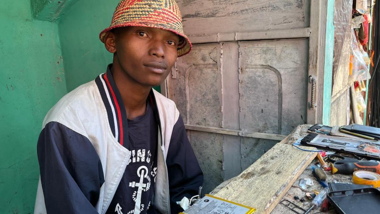Laza Brenda, a young man in a straw hat, sitting at his mobile repair kiosk.