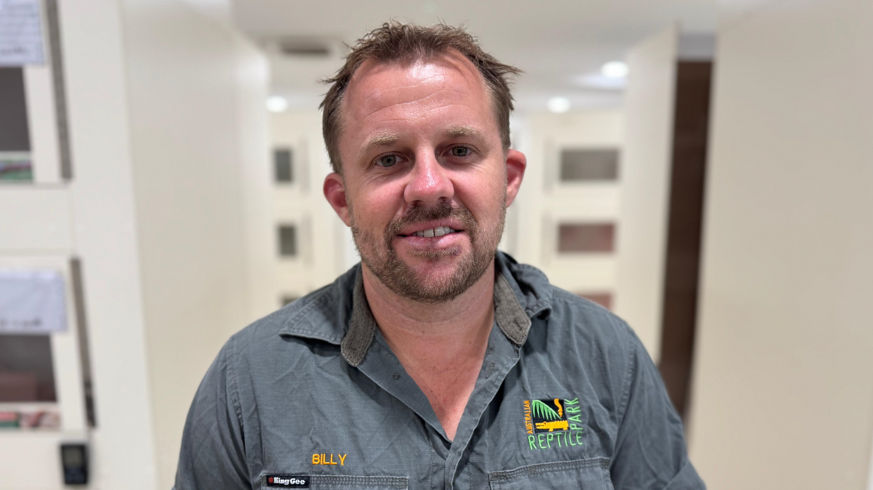 Man smiling and looking directly at camera, wearing a dark grey collared shirt with an Australian Reptile Park logo on the right and "Billy" on the left