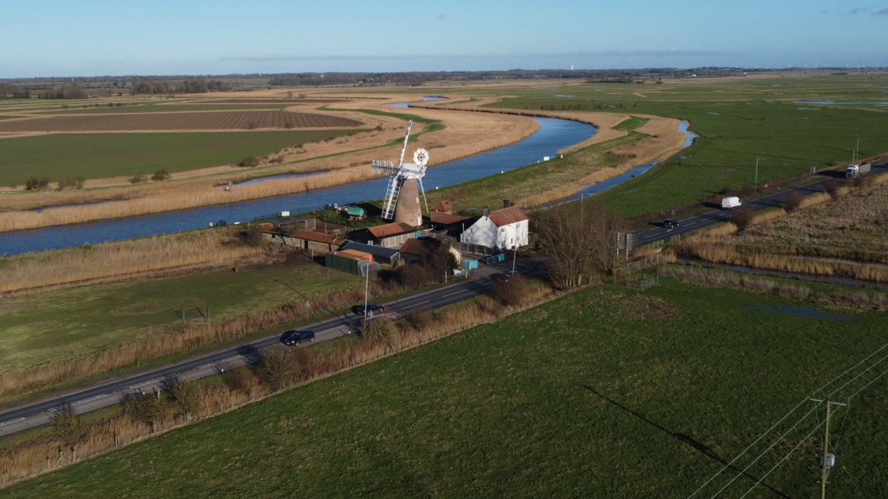 An aerial view of part of the A47 Acle Straight near Great Yarmouth in Norfolk. The road runs between fields, and some houses. a windpump and a river can also be seen.