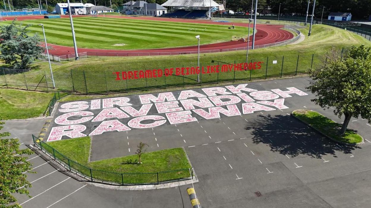 An aerial view of of the white and red mural featuring the names of hundreds of pupils on a stretch of grey tarmac and the red quote which reads "I dreamed of being like my heroes." An orange race track can be seen behind the artwork. A large tree can be seen in the foreground.