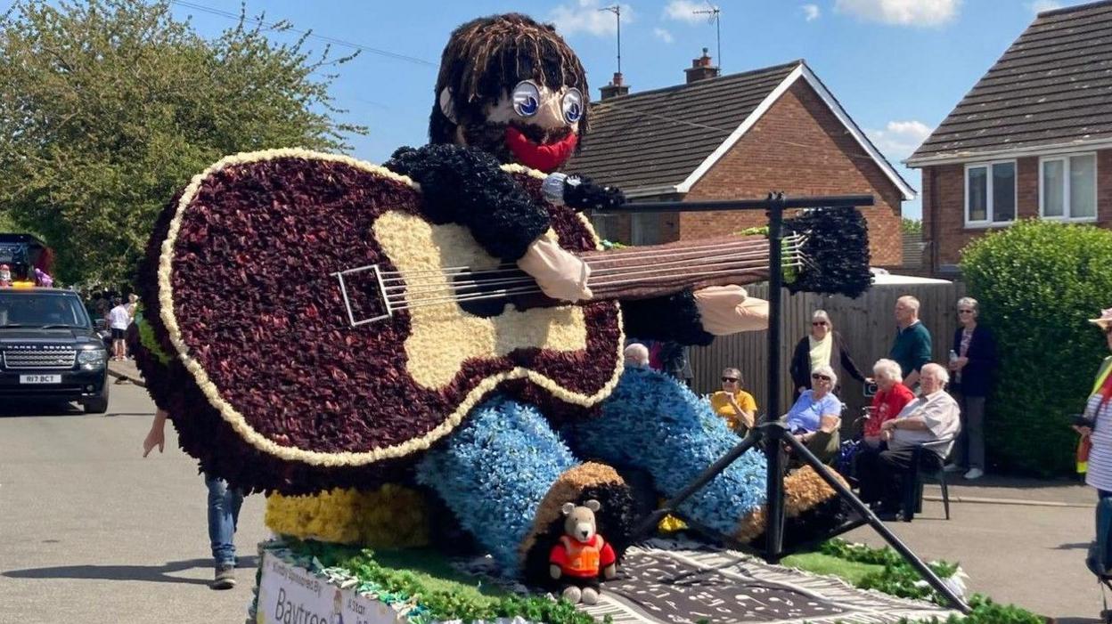 One of the floats is a depiction of a man with a guitar, singing at a microphone. He has brown hair, glasses, red lips and is wearing blue jeans.