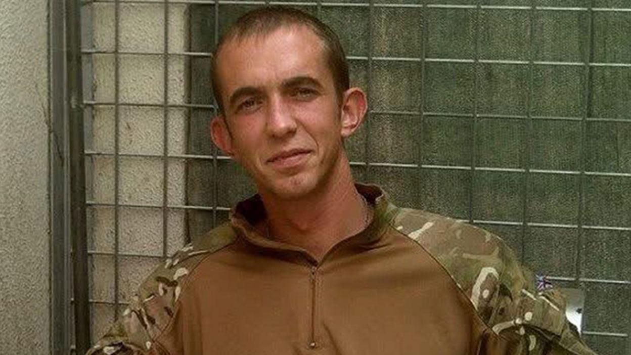 A grainy headshot of Camera Alexander wearing a camouflage Army jacket. He is standing in front of a metal fence and looking into the camera.