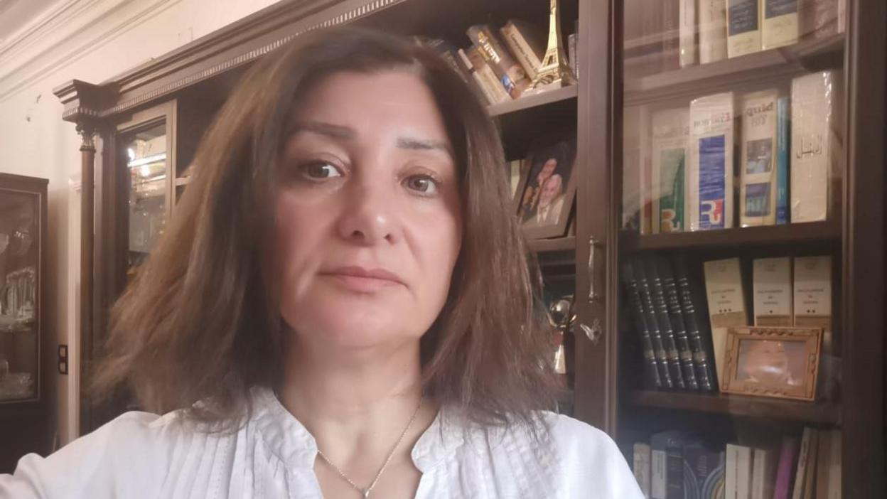 A woman with shoulder length brown hair and in a white shirt looks to camera with a book case behind her