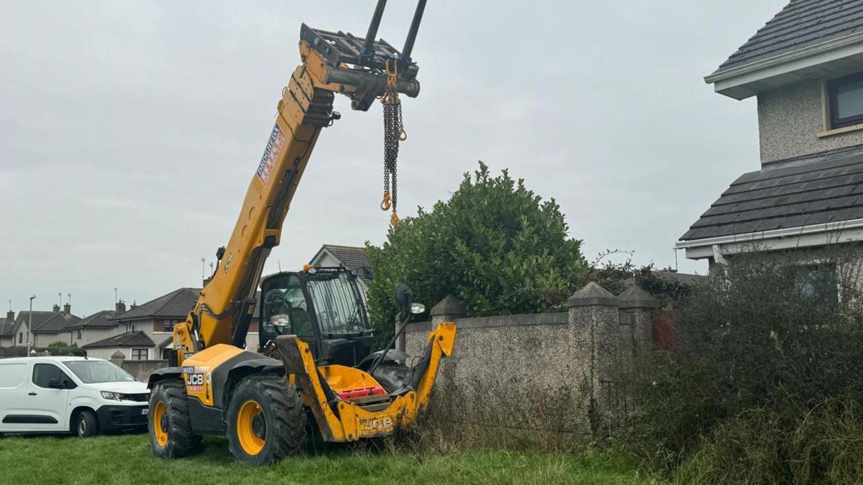 A JCB and a white van parked on a patch of grass. There is a grey wall behind the JCB and homes in the background. 