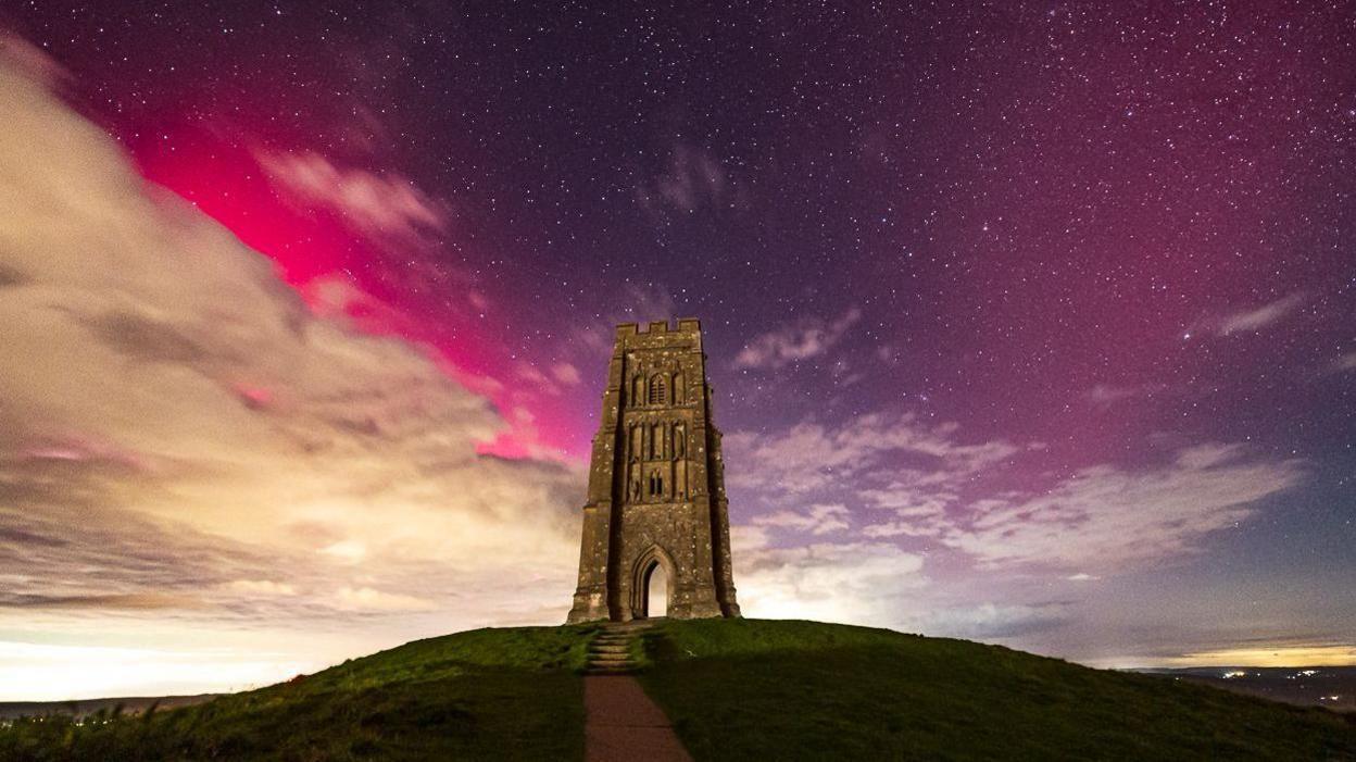 Glastonbury Tor lit up from below. In the sky behind it, pink and purple lights are flooding the sky. There are also thousands of dotted stars and light clouds covering the left of the picture. 