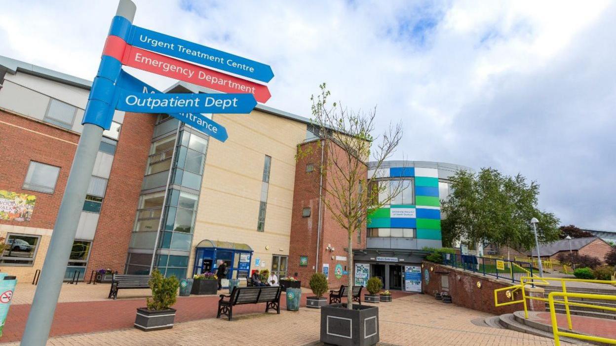 The outside of the University of North Durham Hospital. It a brown and beige-coloured brick building. The entrance has green, white and blue rectangle panelling. There is a sign pointing patients to various departments.  A small tree is in a plant pot in a seating area.