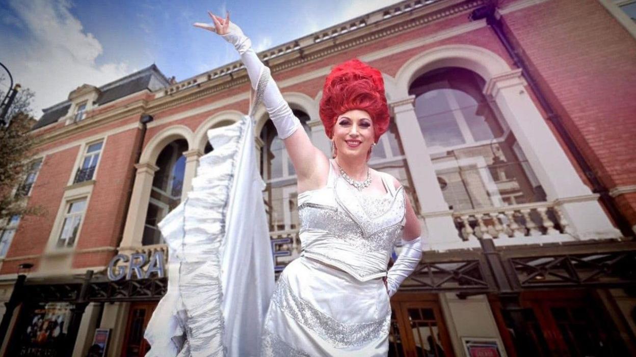 A woman with tall red hair is smiling at the camera standing outside a theatre. She is wearing a white dress and her right arm is outstretched above her head.