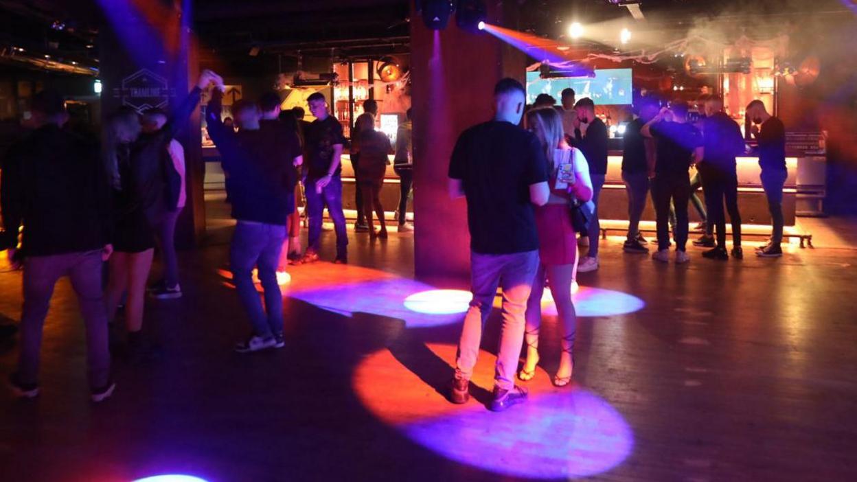 People dancing inside a nightclub. The clubbers are spaced out across the room, some are dancing and some are talking.  Multi-coloured lights are lighting up the dark room.